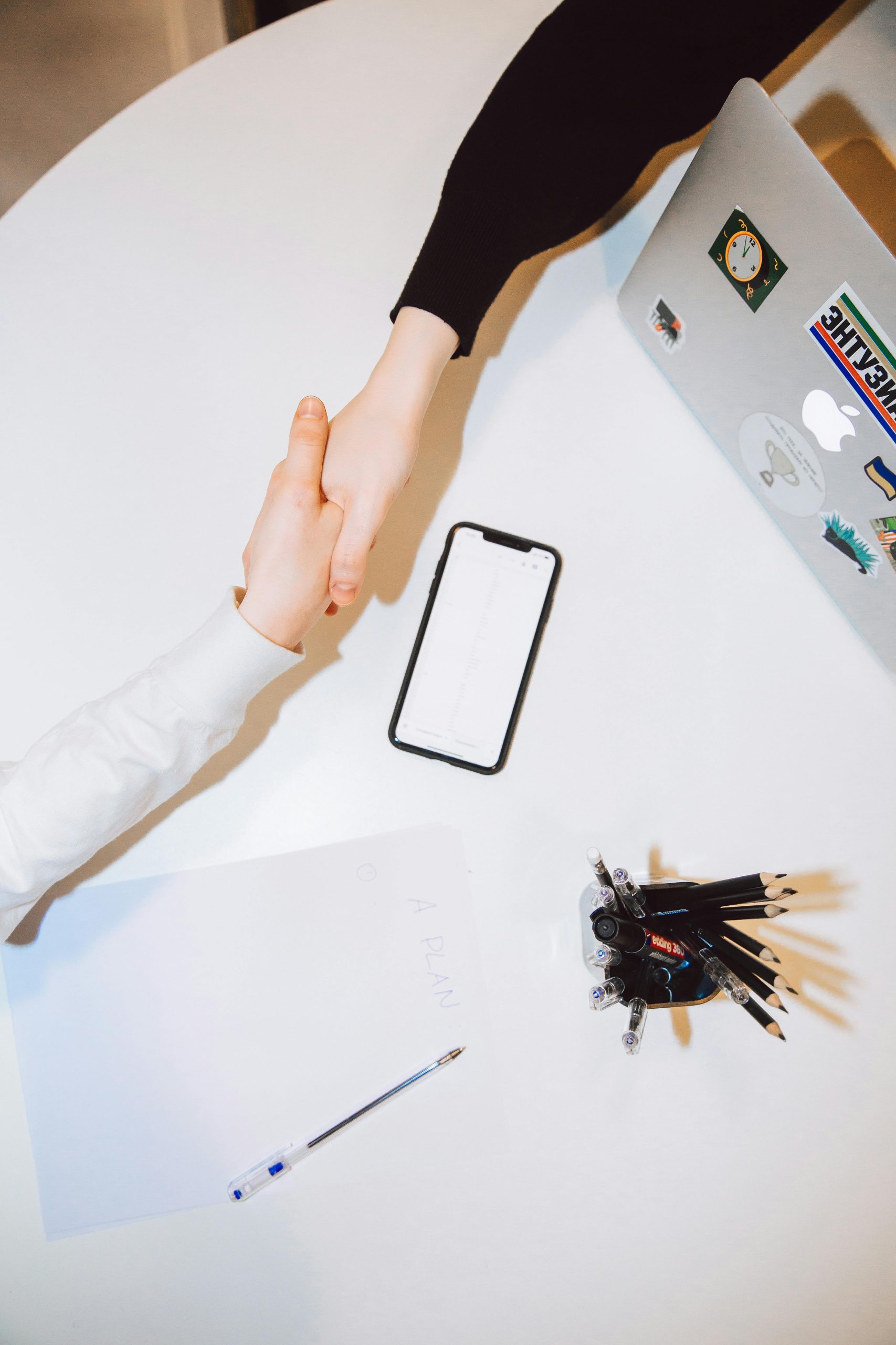 Inicio Two individuals shaking hands across a desk with a smartphone and laptop in a modern office setting.